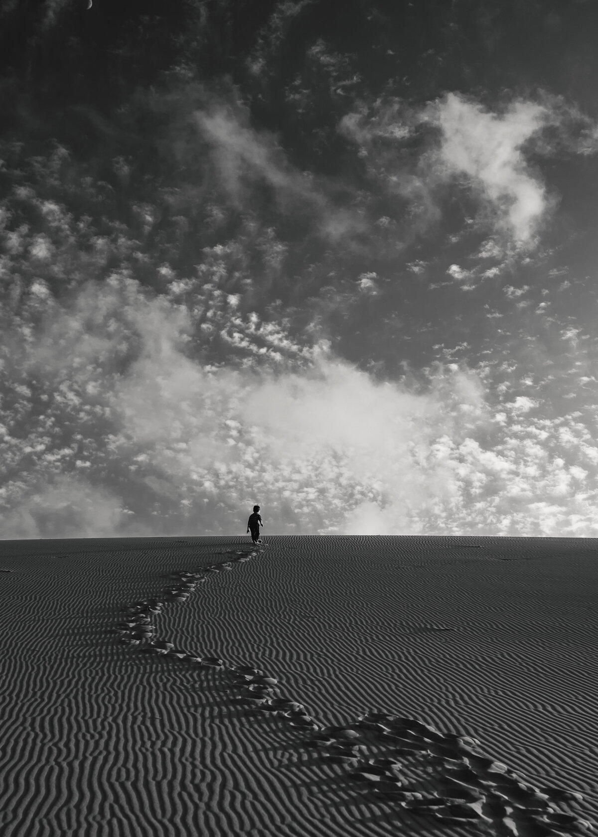 Environmental portrait of boy walking up sand dune toward cloudy sky