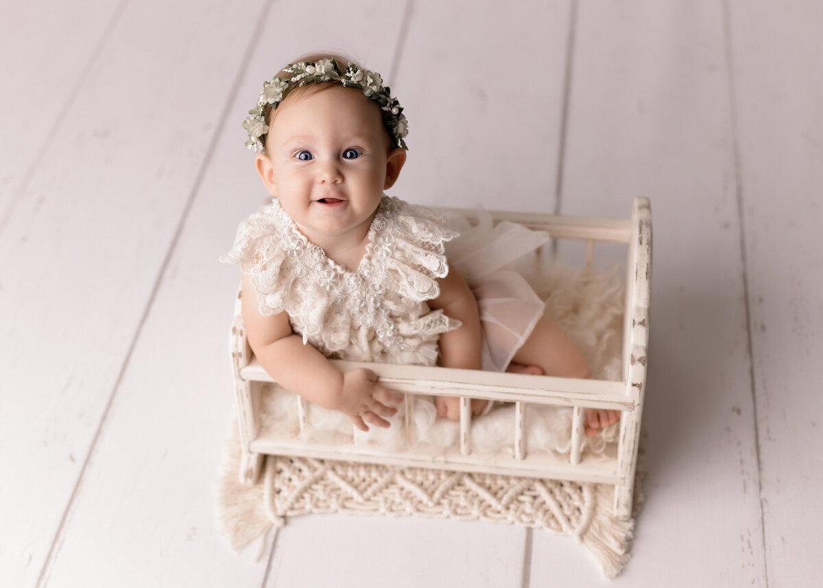 12-month baby photos at top West Palm Beach and Boynton Beach, FL photography studio. Baby in a vintage miniature doll crib. She is wearing a cream lace dress and floral crown with her arm draped over the edge of the crib. Baby is smiling up at the camera.