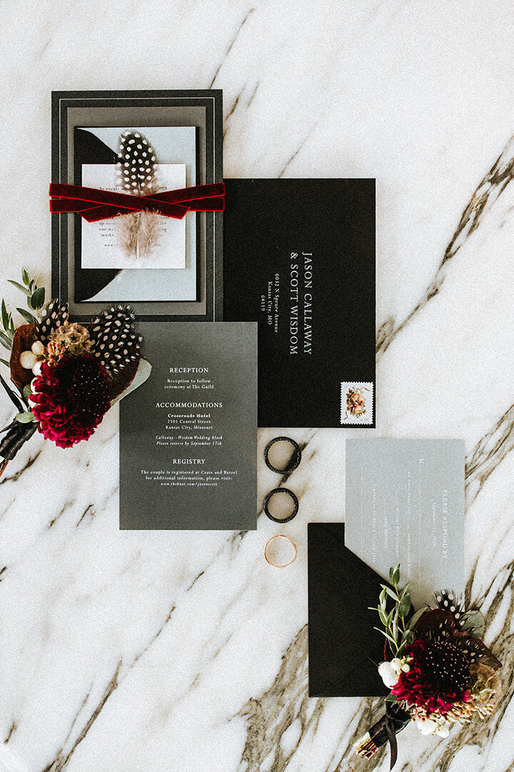 Various black and gray wedding stationery with white lettering atop a white and gold marble background.