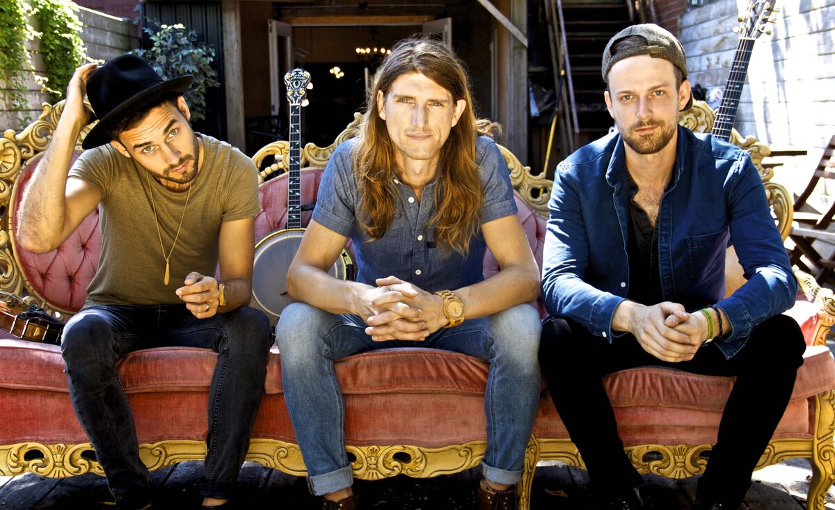Musical trio portrait The East Pointers sitting against pink sofa member tipping black fedora other members hands clasped together
