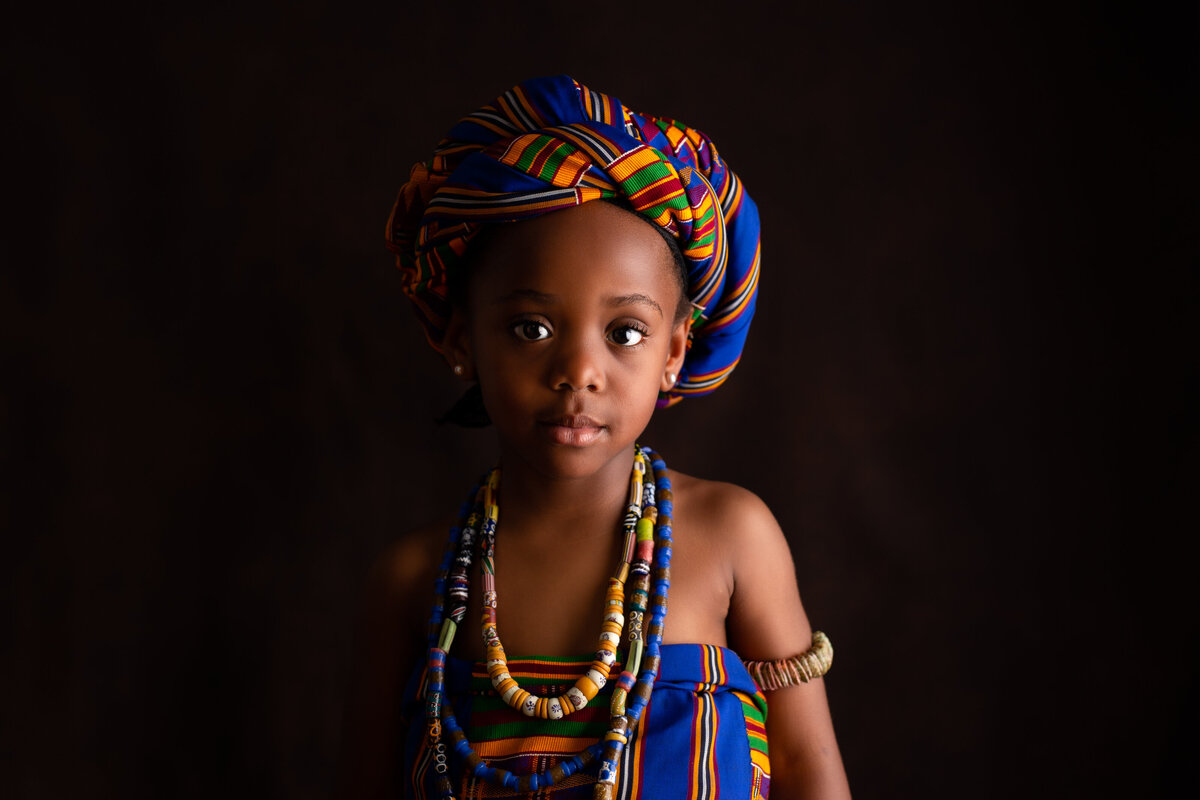 Little girl in colouful traditional dress wearing a head wrap and beads around her neck on a black background.