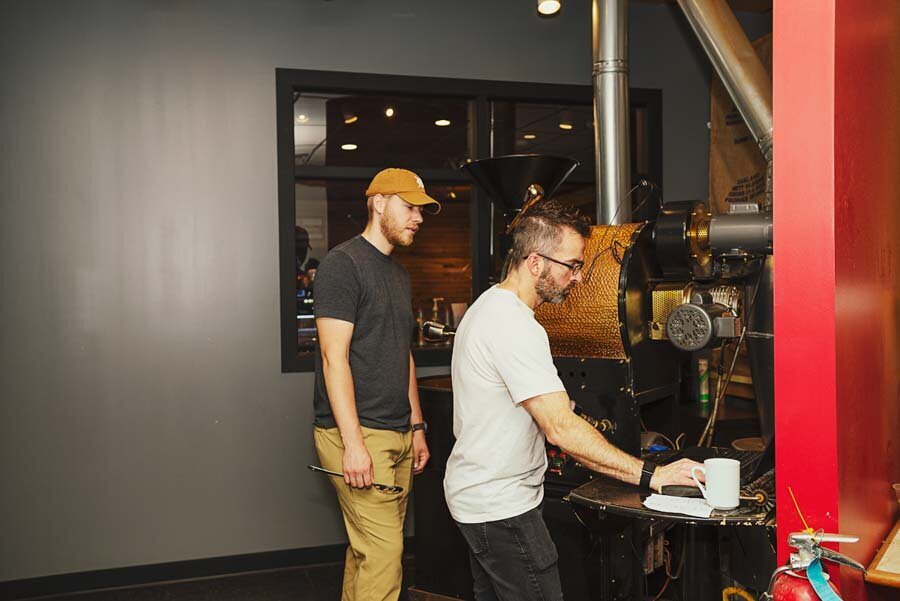 Two men working with a coffee roaster in a cafe. one observes while the other handles the machine.