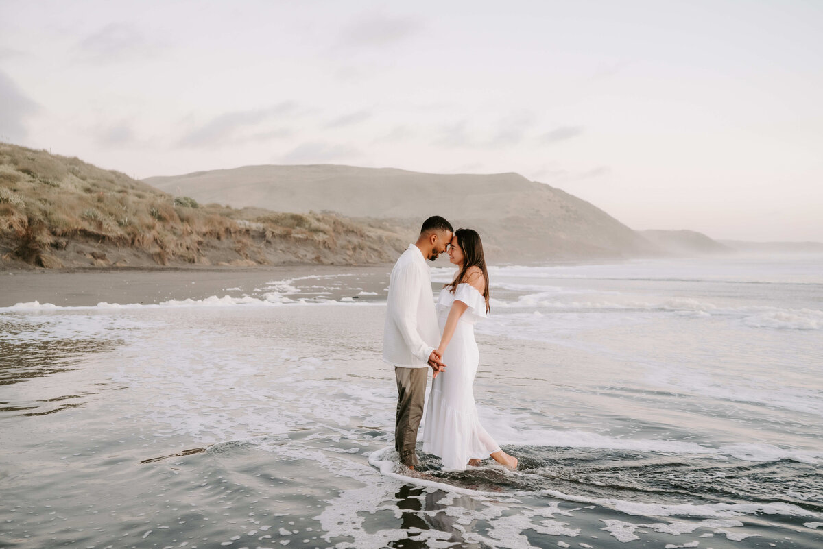 Engaged couple standing in the ocean at sunset, holding hands and touching foreheads, dressed in white for a romantic beach photoshoot.