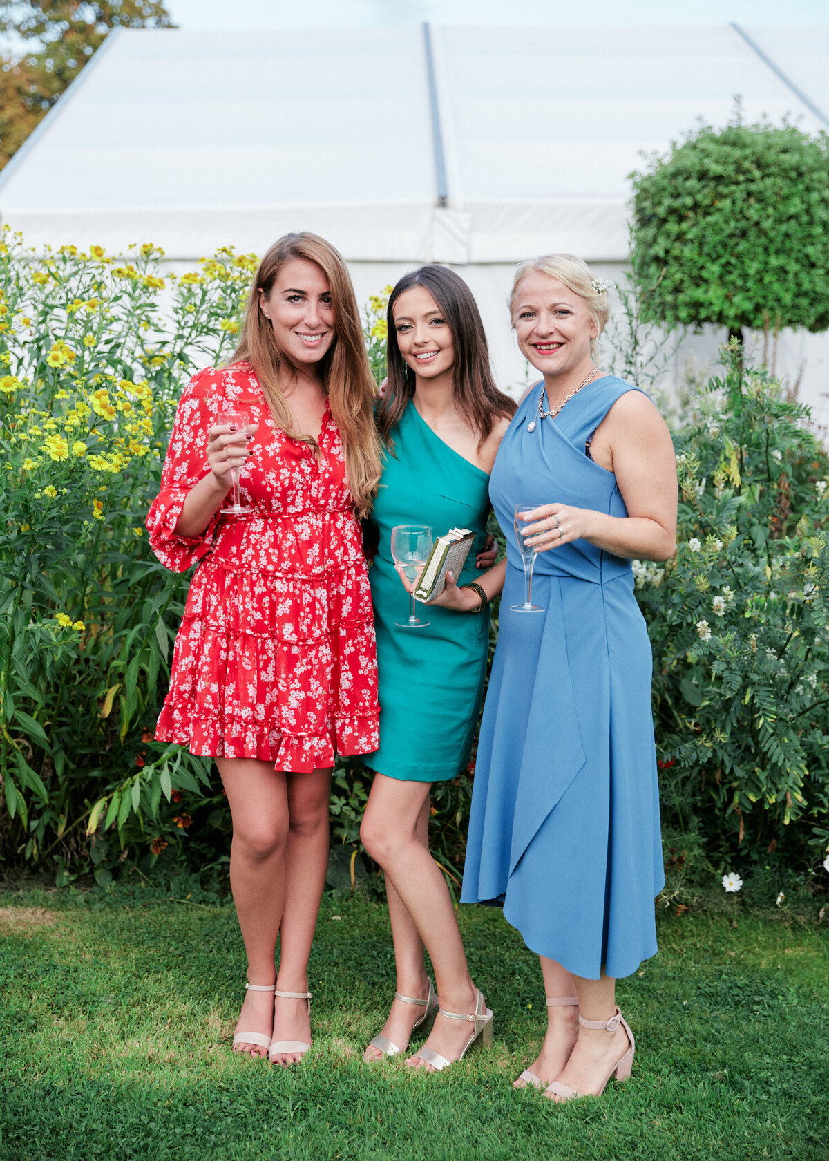 three girls wearing colourful red green and blue dresses holding champagne glasses and posing for photo