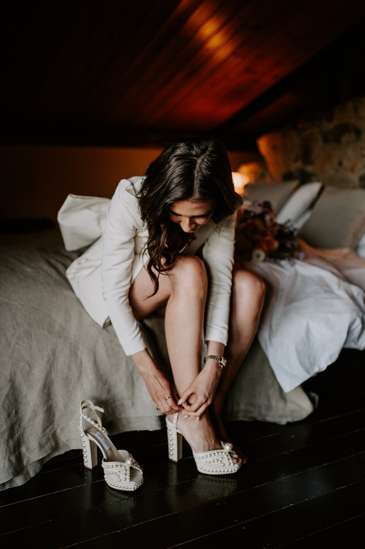 A bride changes from her fringed wedding dress into this shorter styled dress with long sleeves and a bow on the back She is sitting on a bed fastening her Jimmy Choo heels.