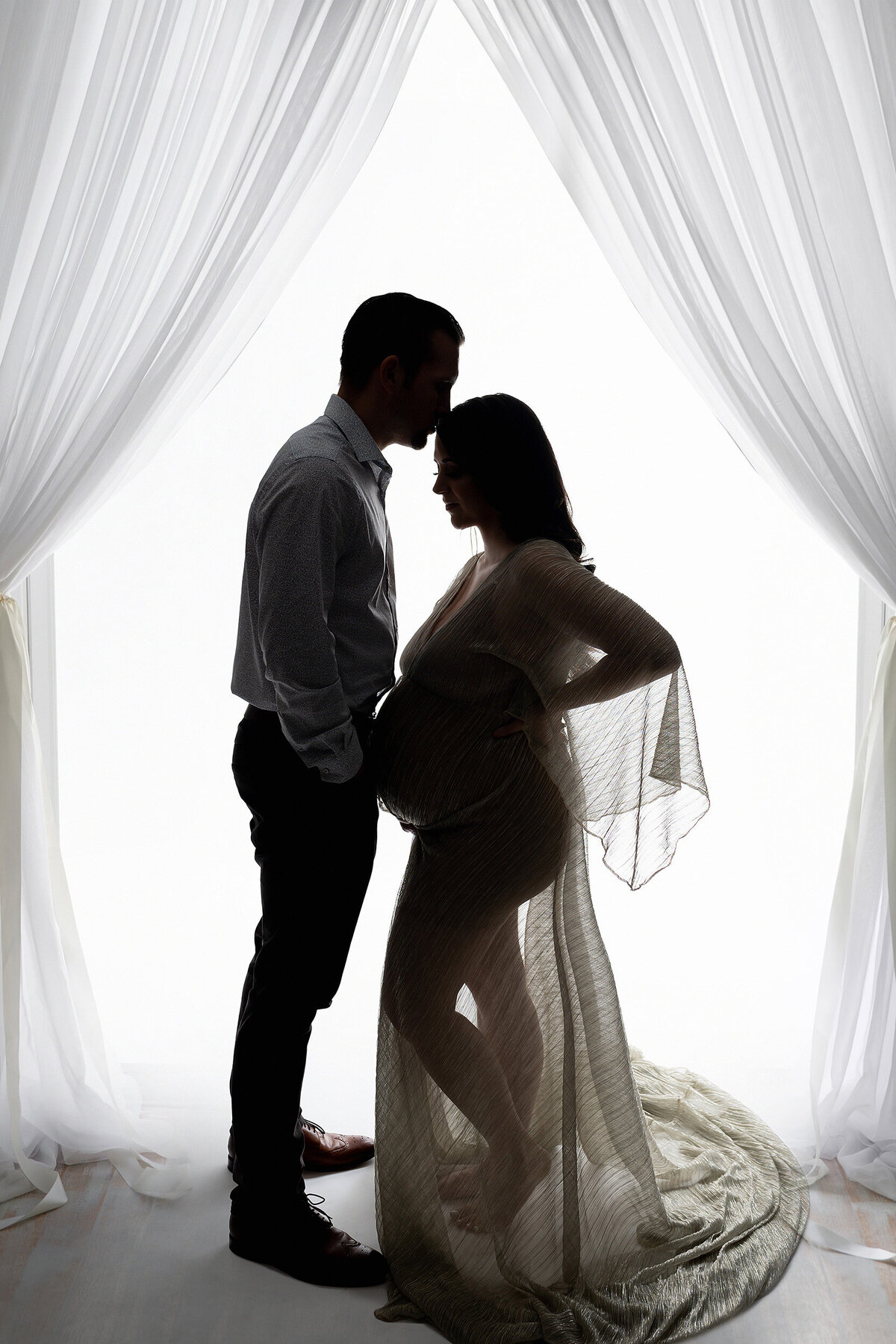 Couple facing eachother shown in shadow with white-draped window behind. Woman cradles her pregnant belly while partner kisses her forehead.