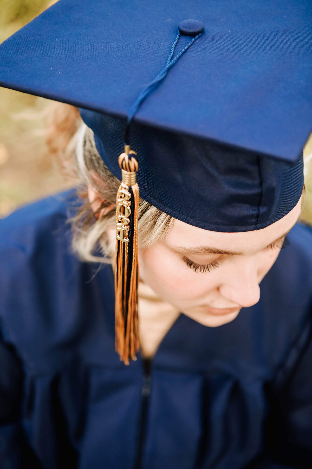 A high school senior in dark blue cap and gown looks down. The picture is taken from above so you can see the gold and blue tassell on her cap
