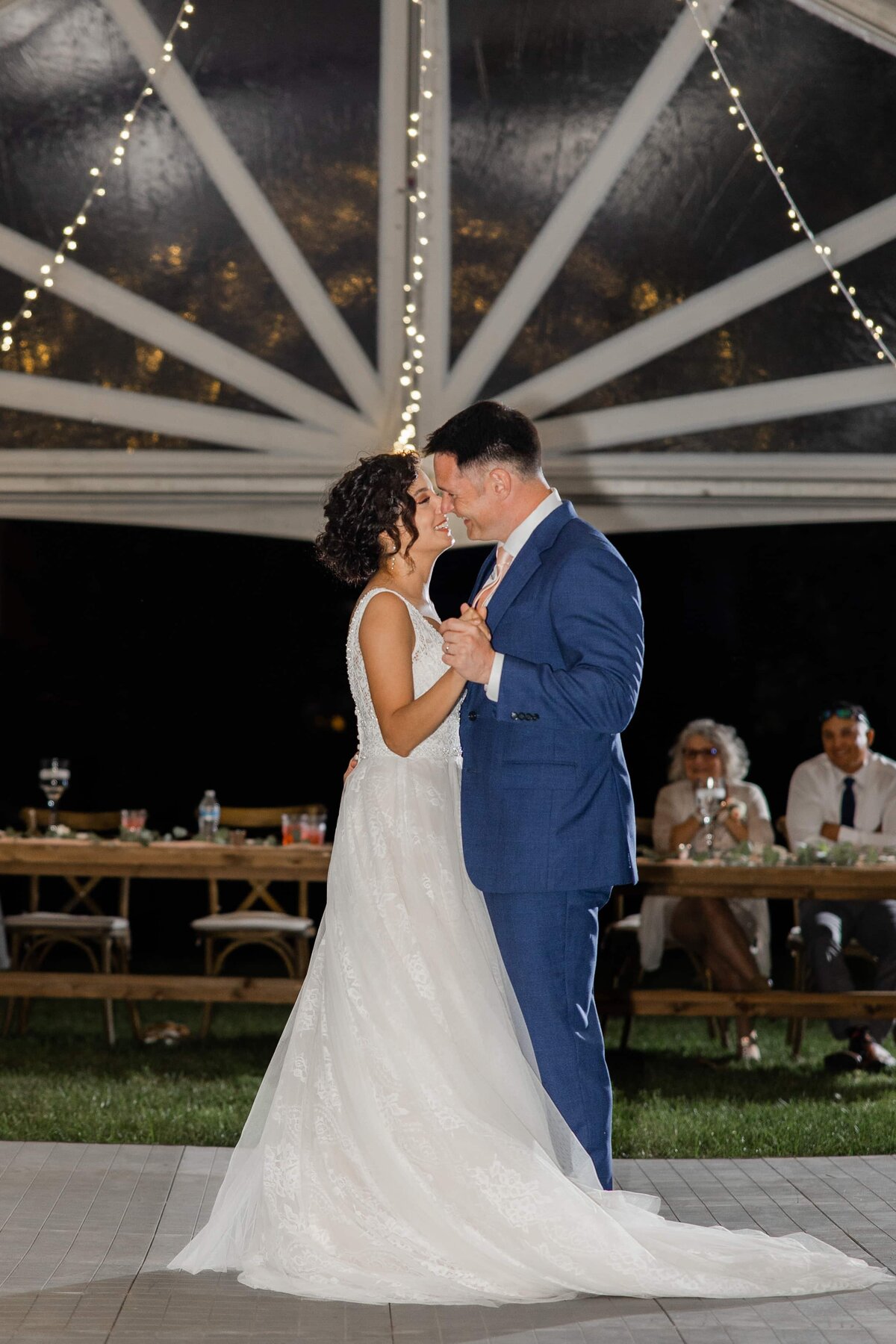 A bride and groom share a romantic dance under a tent with string lights at an Iowa wedding, with guests watching in the background.
