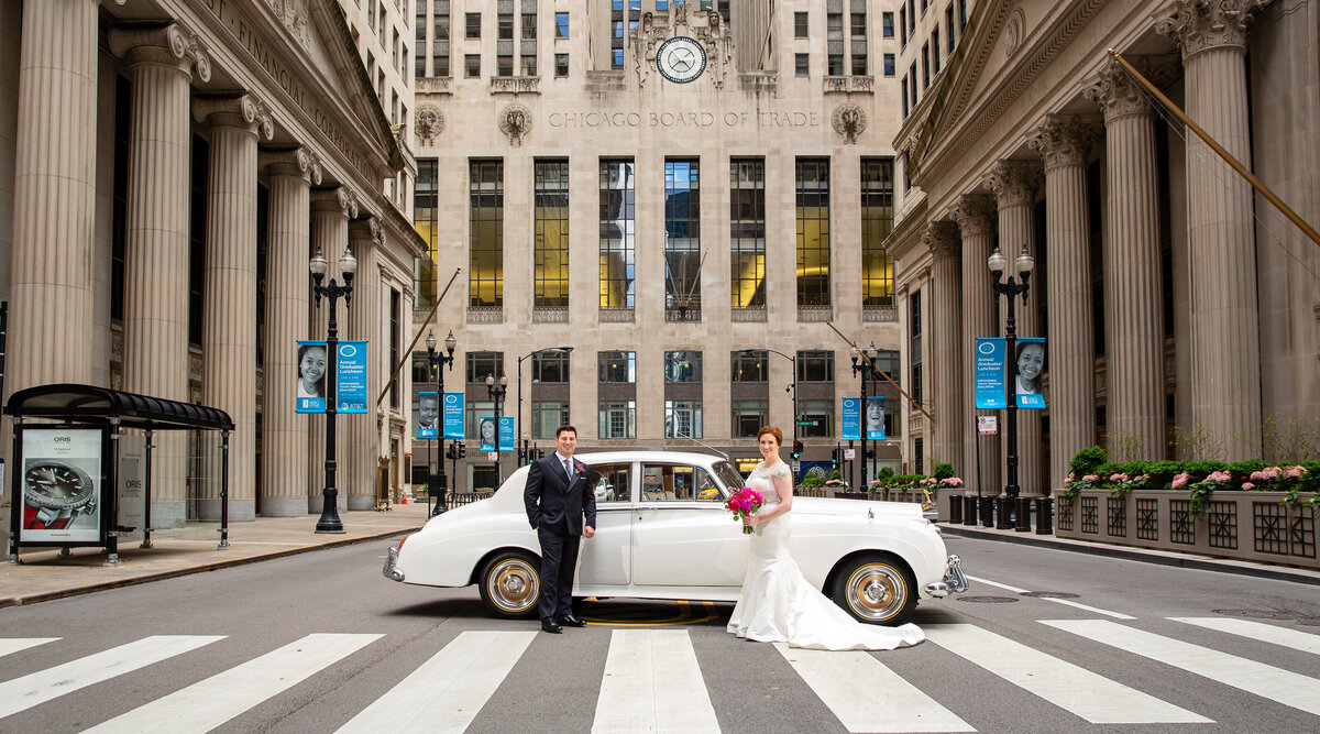 A bride and groom with a vintage car parked in the middle of the street at the Chicago Board of Trade building.