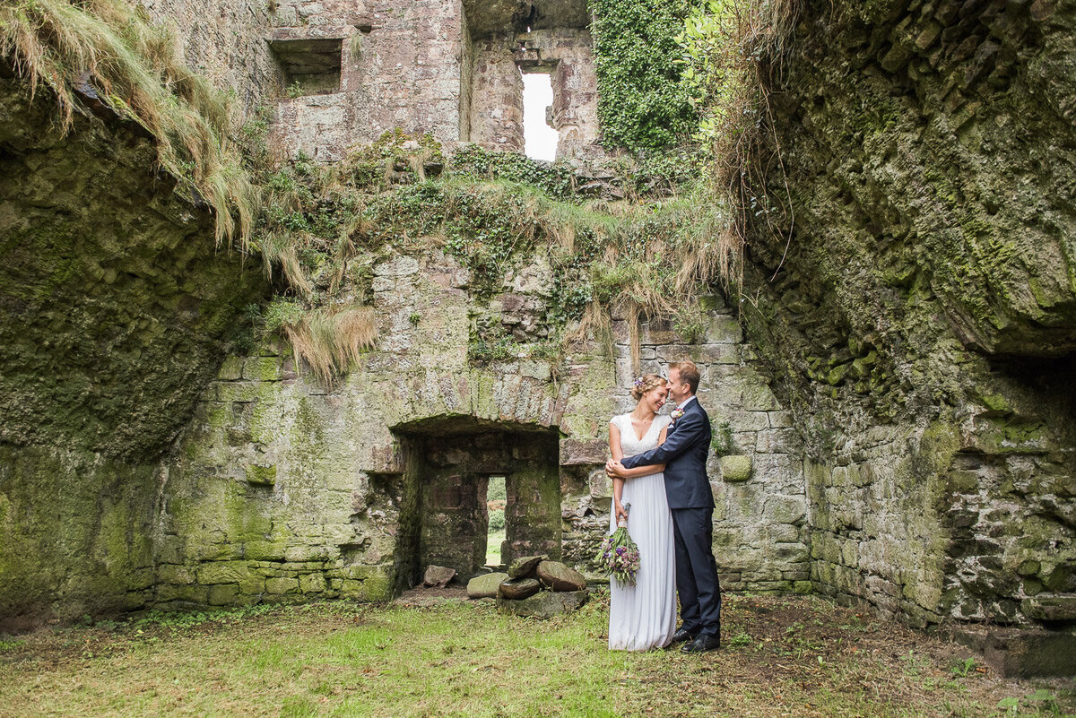 bride wearing a beaded column style wedding dress with sleeves, holding a wild flower bouquet, standing with her husband in a navy suit in the ruins of Minard Castle, West Kerry