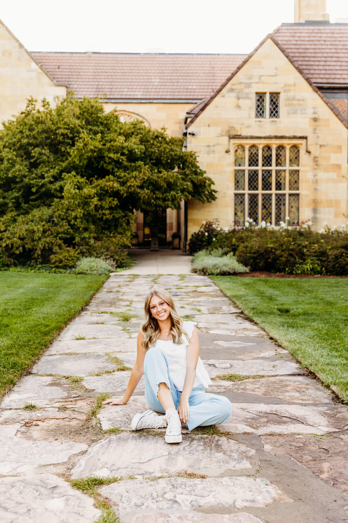 pretty teen girl in a white top and denim bottoms sitting with one knee up and the other crossed below on a stone pathway and smiling  captured by Green Bay senior photographer Ashley Kalbus
