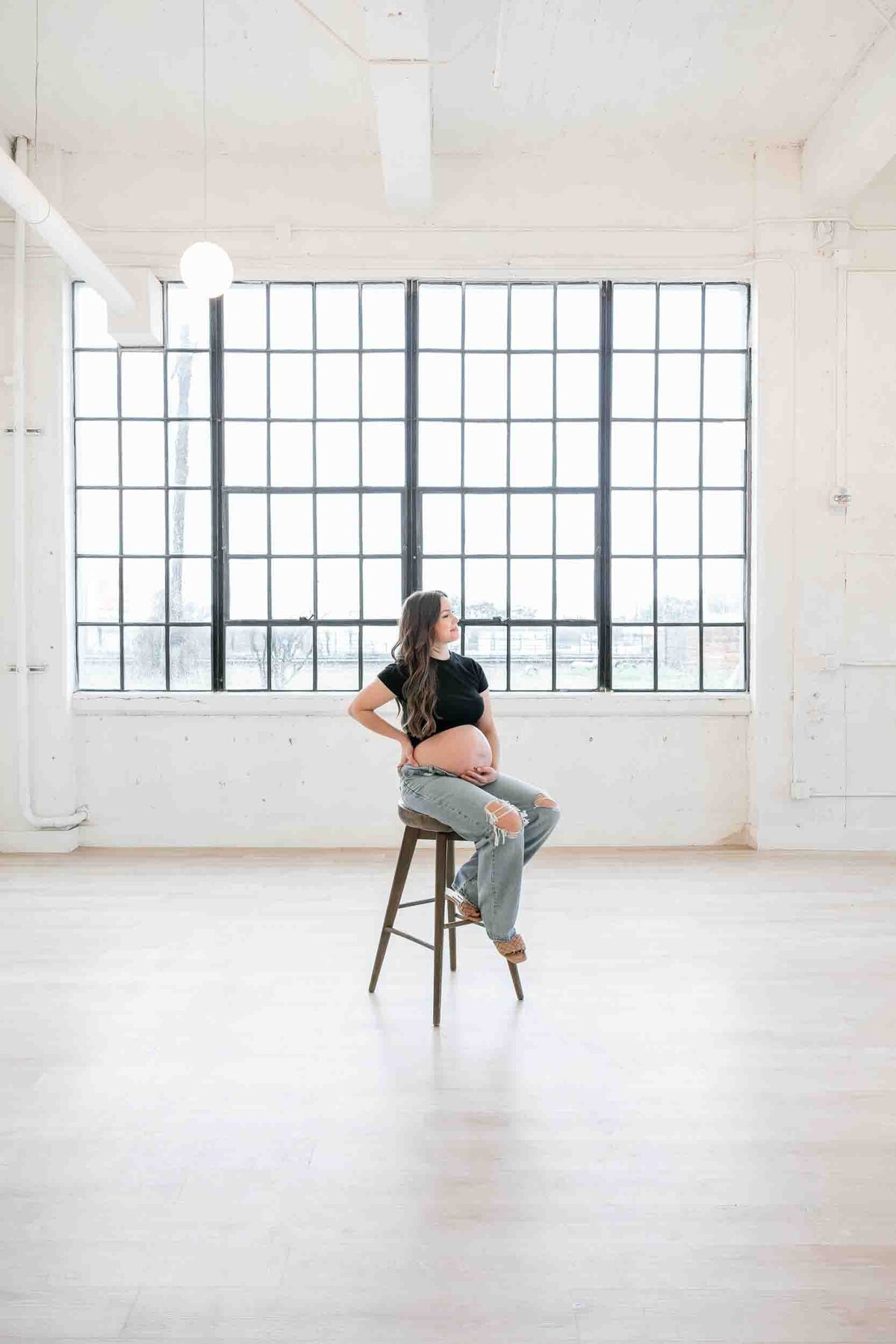 pregnant woman sits on stool, holding her bare pregnant belly, in brightly lit studio.