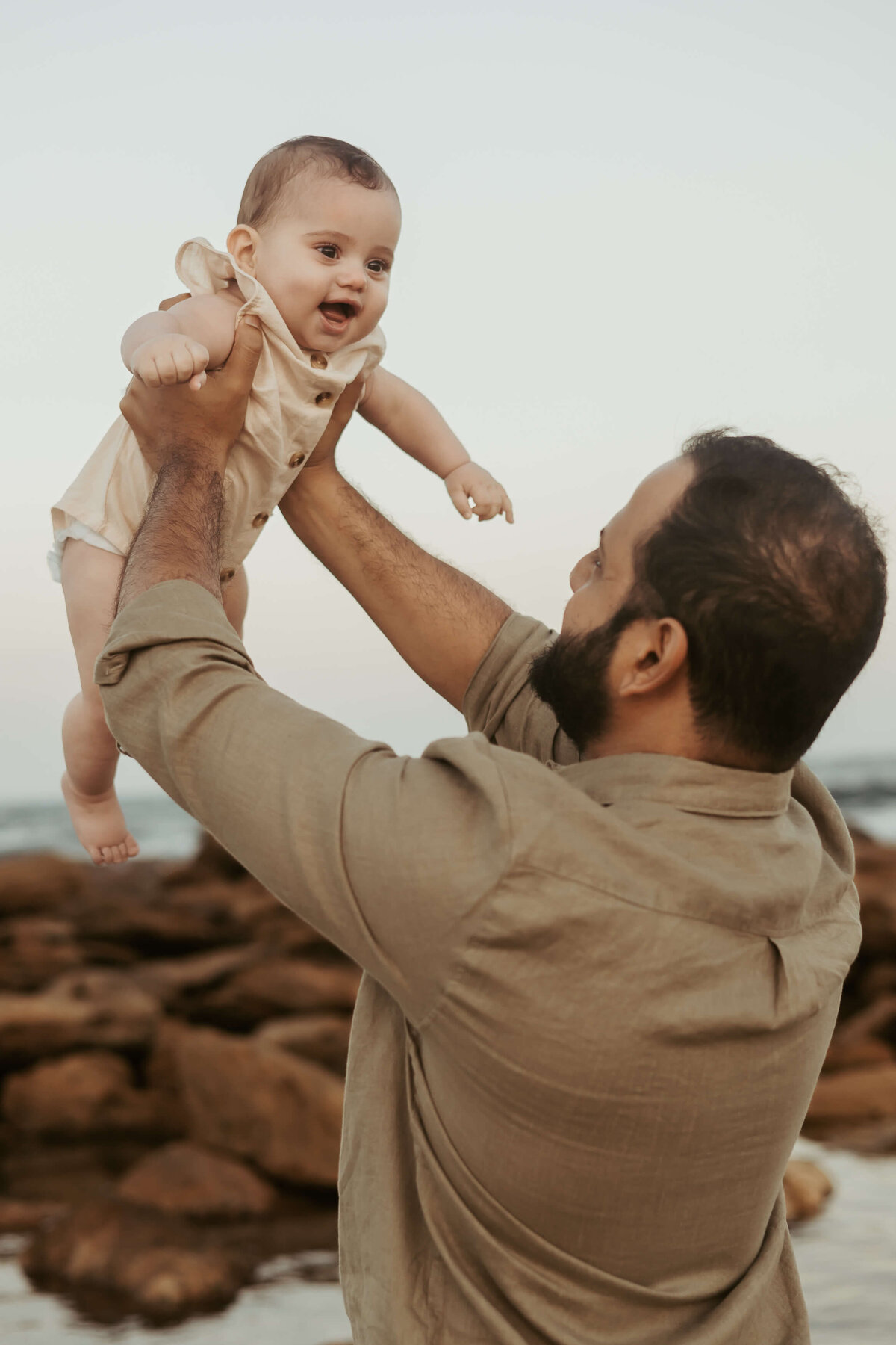 Dad has his back to the camera as he holds up his giggling baby during a beautiful beach sunset.