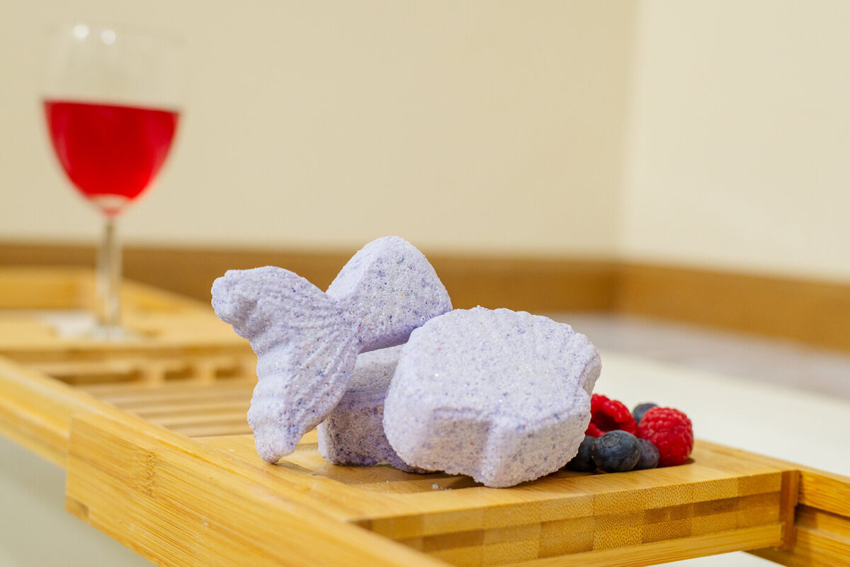 Purple butterfly-shaped and round bath bombs on a wooden tray next to fresh berries and a glass of red wine.