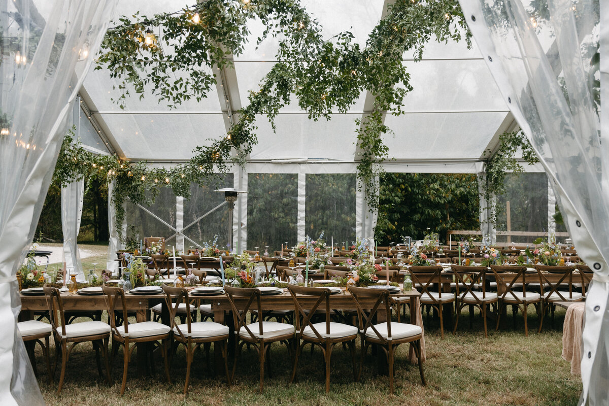 Long wooden reception tables arranged under a greenery-draped clear tent with pastel floral centerpieces, taper candles, and vintage glassware at The Nest wedding venue in Northwest Arkansas.
