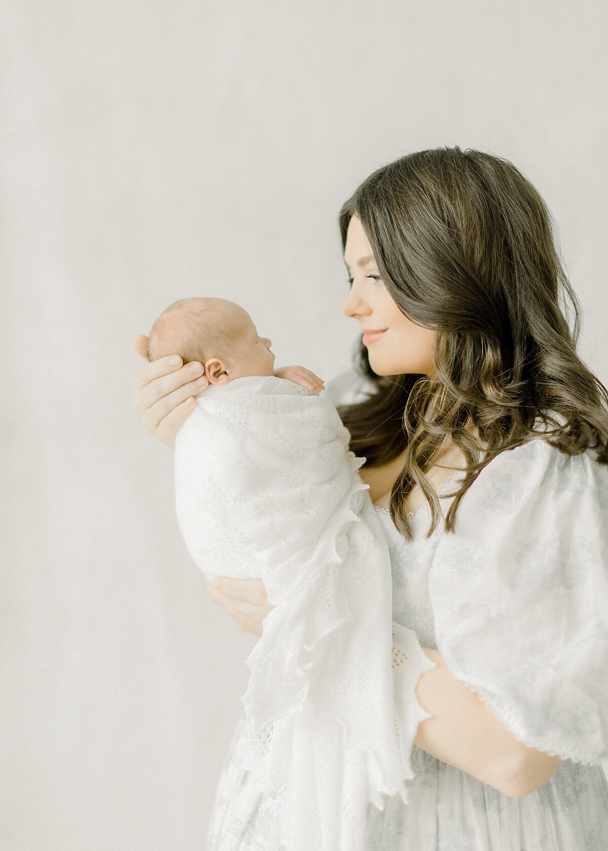 A mother standing in a Dallas Texas photography studio holding her newborn baby boy up close as she smiles and embraces him.