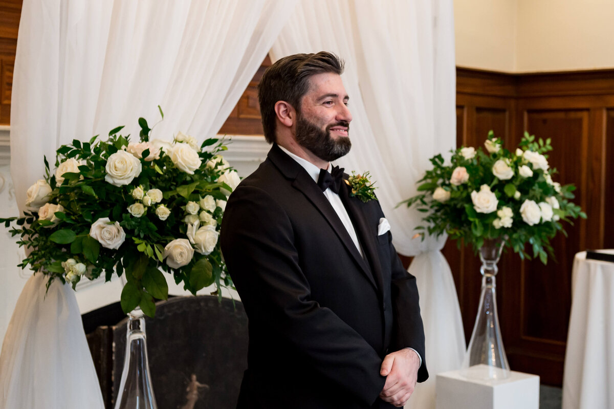 a groom in a black tuxedo smiling as he sees his bride approach during his Ottawa wedding ceremony at the Chateau Laurier hotel