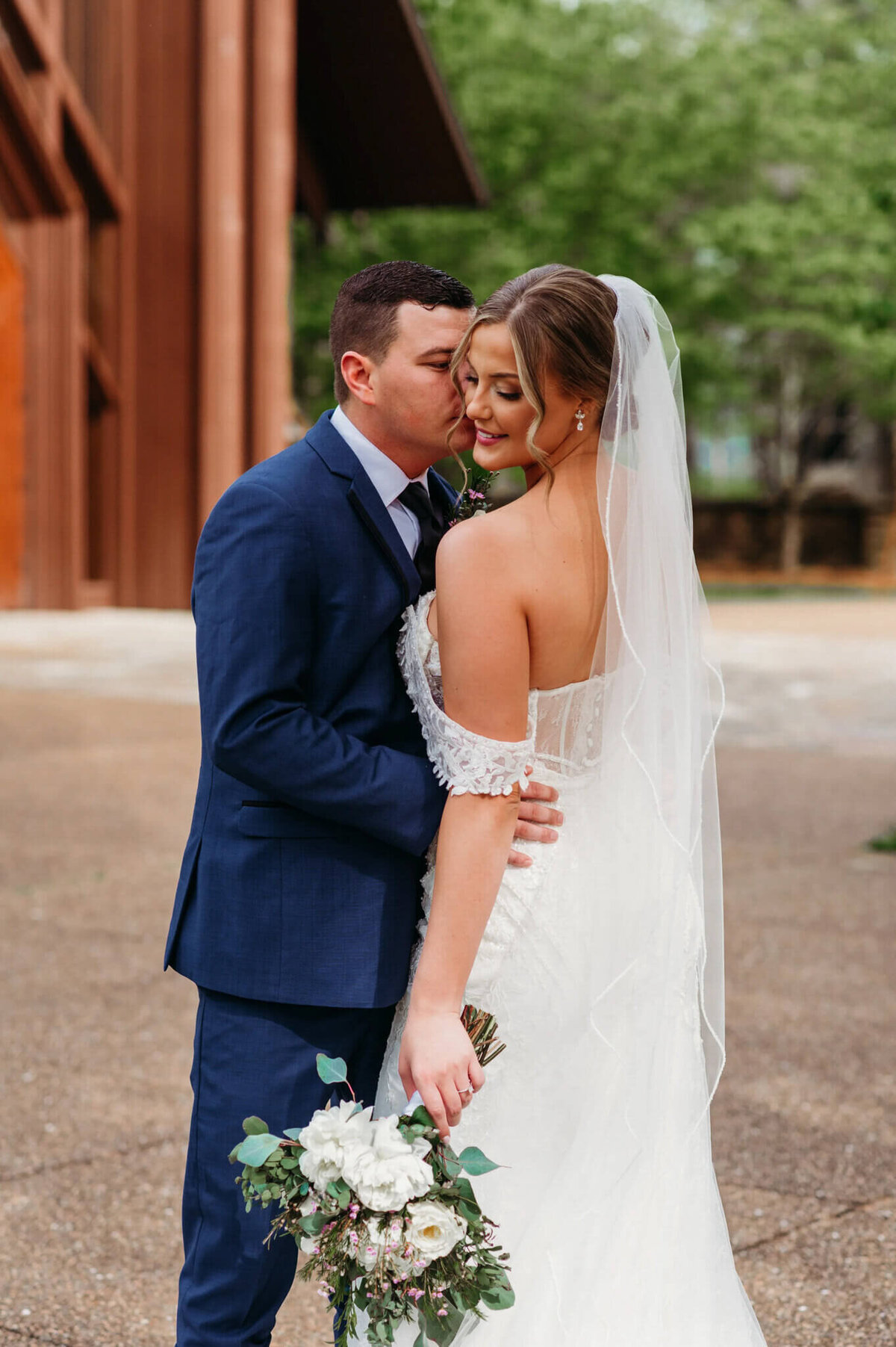 Photo of a groom kissing his bride cheek while she looks at the ground and smiles