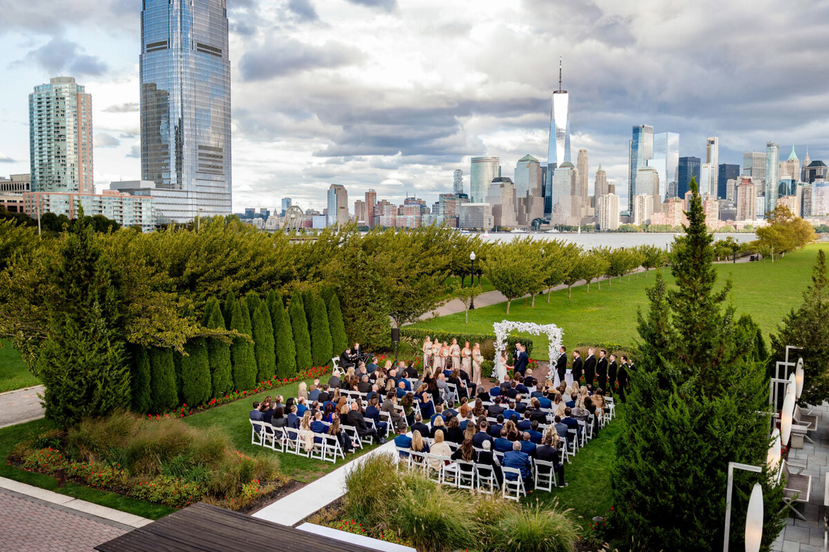 An aerial view of a wedding ceremony, taking place outside of liberty House with the New York City skyline in the background