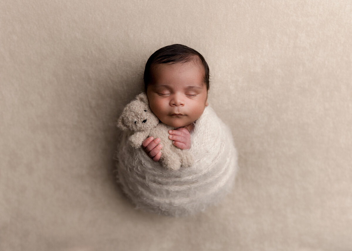 Newborn studio portrait by top West Palm Beach photographer, Michelle Lee Photography. Newborn baby boy is swaddled in a cream-colored swaddle with his fingers peeking out of the top holding a petite beige teddy bear. Baby is sleeping with the teddy bear resting on his cheek.