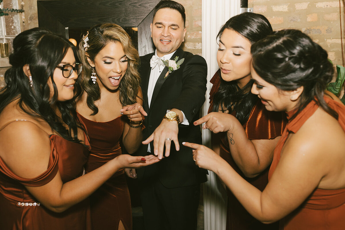 Four bridesmaids in elegant red gowns stand around Chicago groom and mockingly stare at wedding band.