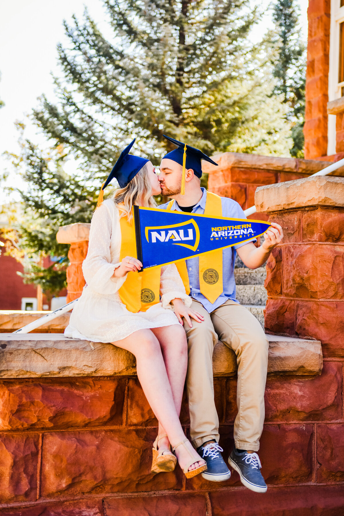 NAU couple kissing at Old Main holding banner sitting on steps in graduation attire