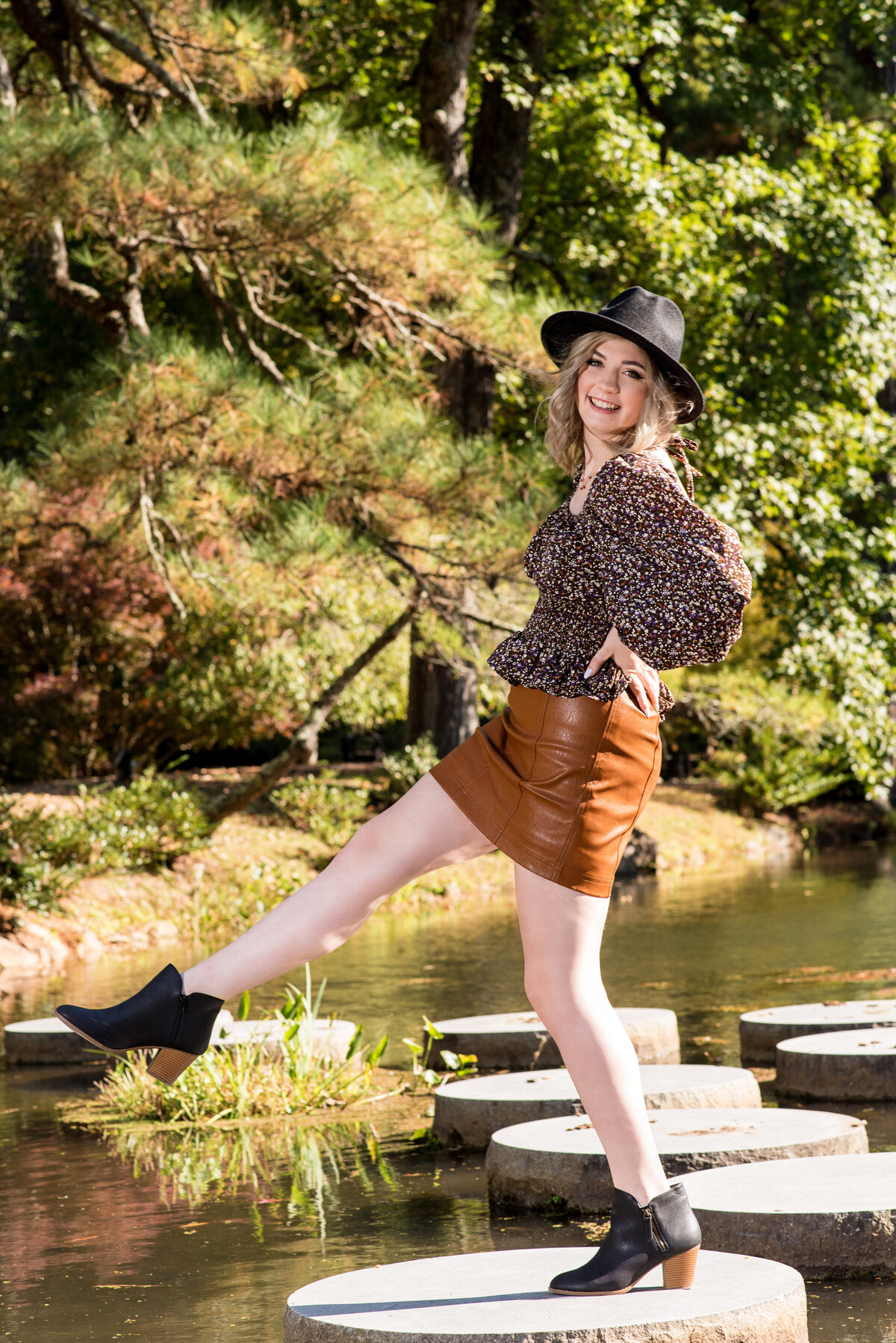 Warsaw high school senior girl wearing floral top and brown leather skirt kicks out leg during Richmond senior portrait shoot at Maymond.