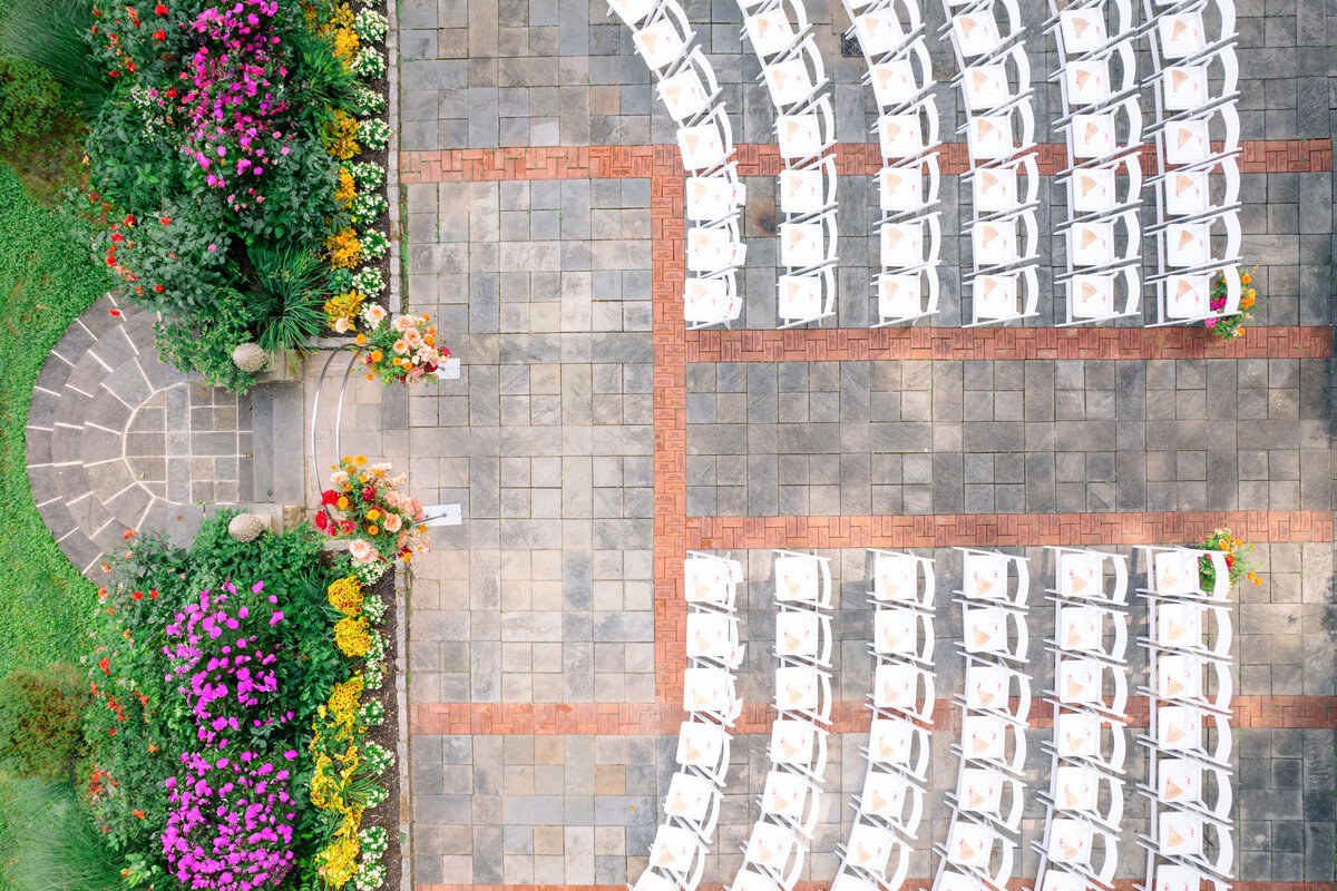 An aerial view of a garden wedding setup with rows of white chairs arranged on a paved surface surrounded by colorful flowers and greenery. A small arch of yellow and orange flowers is at the front.
