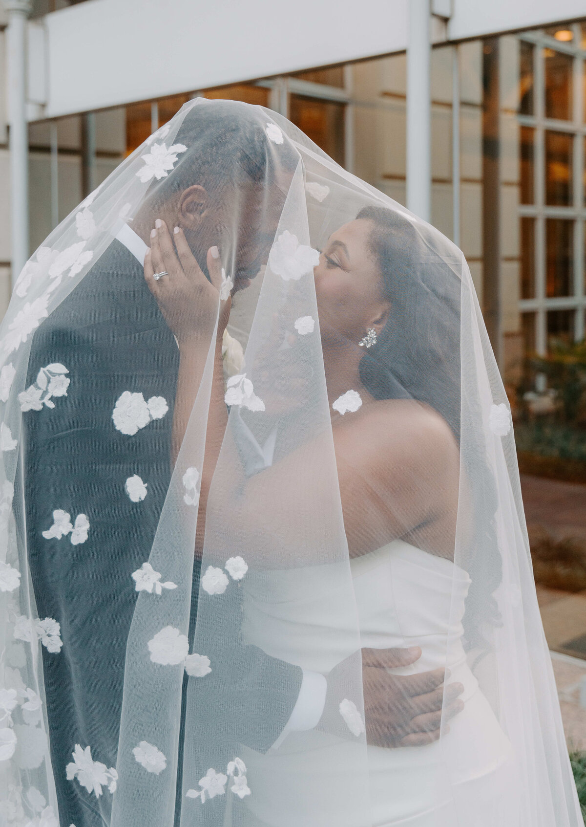 Bride and groom share a tender kiss under a flower-adorned veil. The bride's hand rests on the groom's cheek, creating a romantic and intimate scene.
