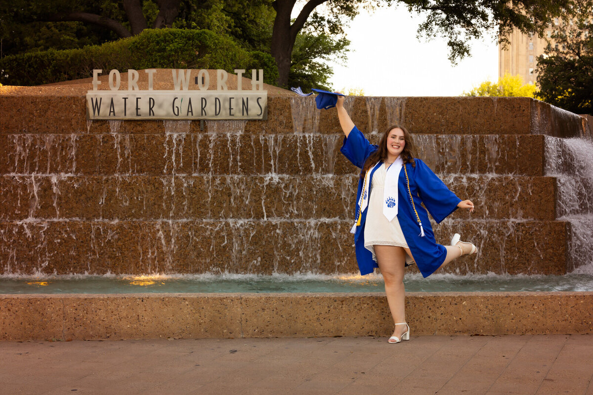 senior-girl-wearing-cap-gown-jumping-at-fort-worth-water-gardens