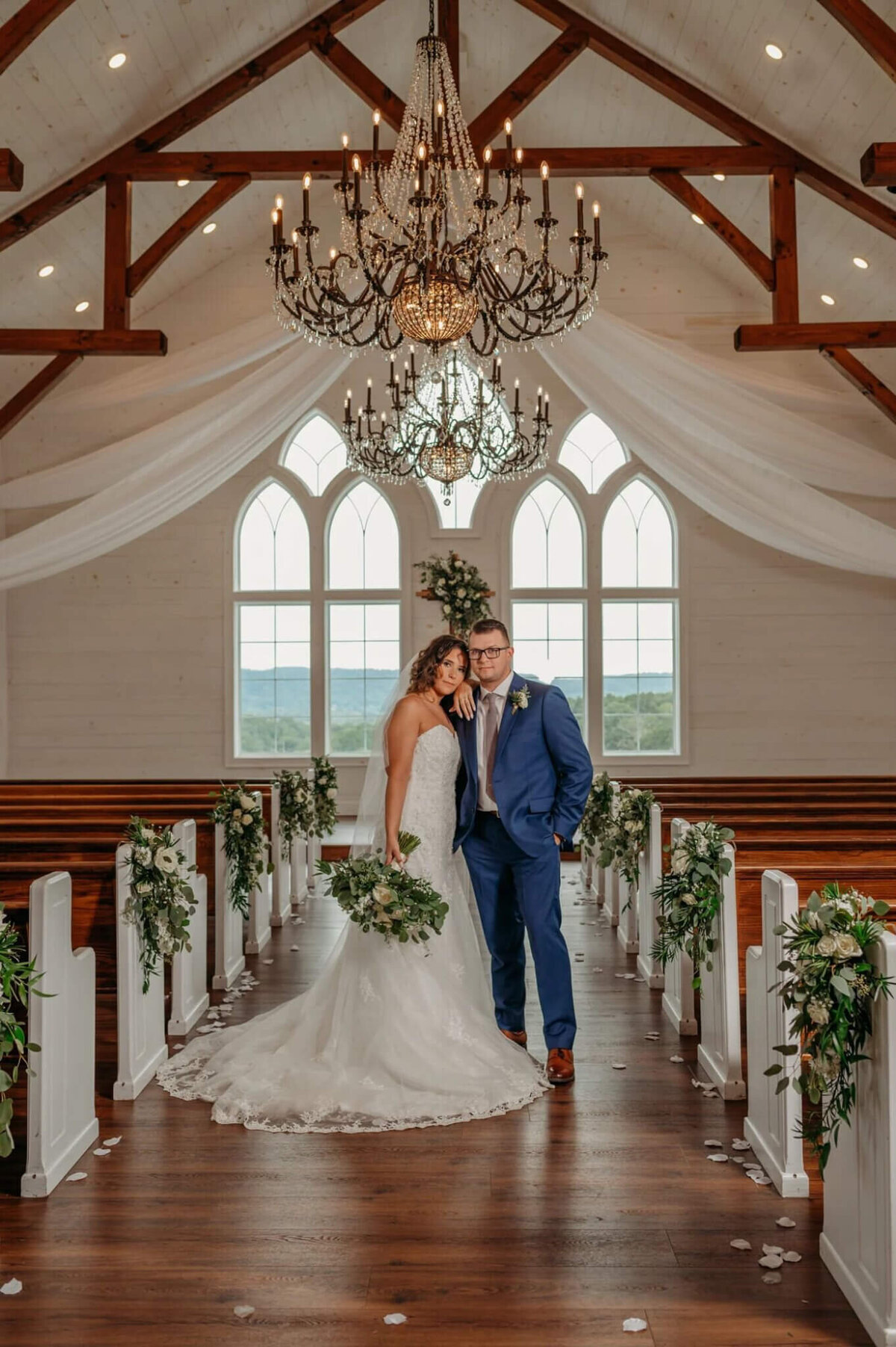 Photo of a bride leaning on her grooms shoulder underneath a chandelier and in between wooden benches of a white wedding chapel