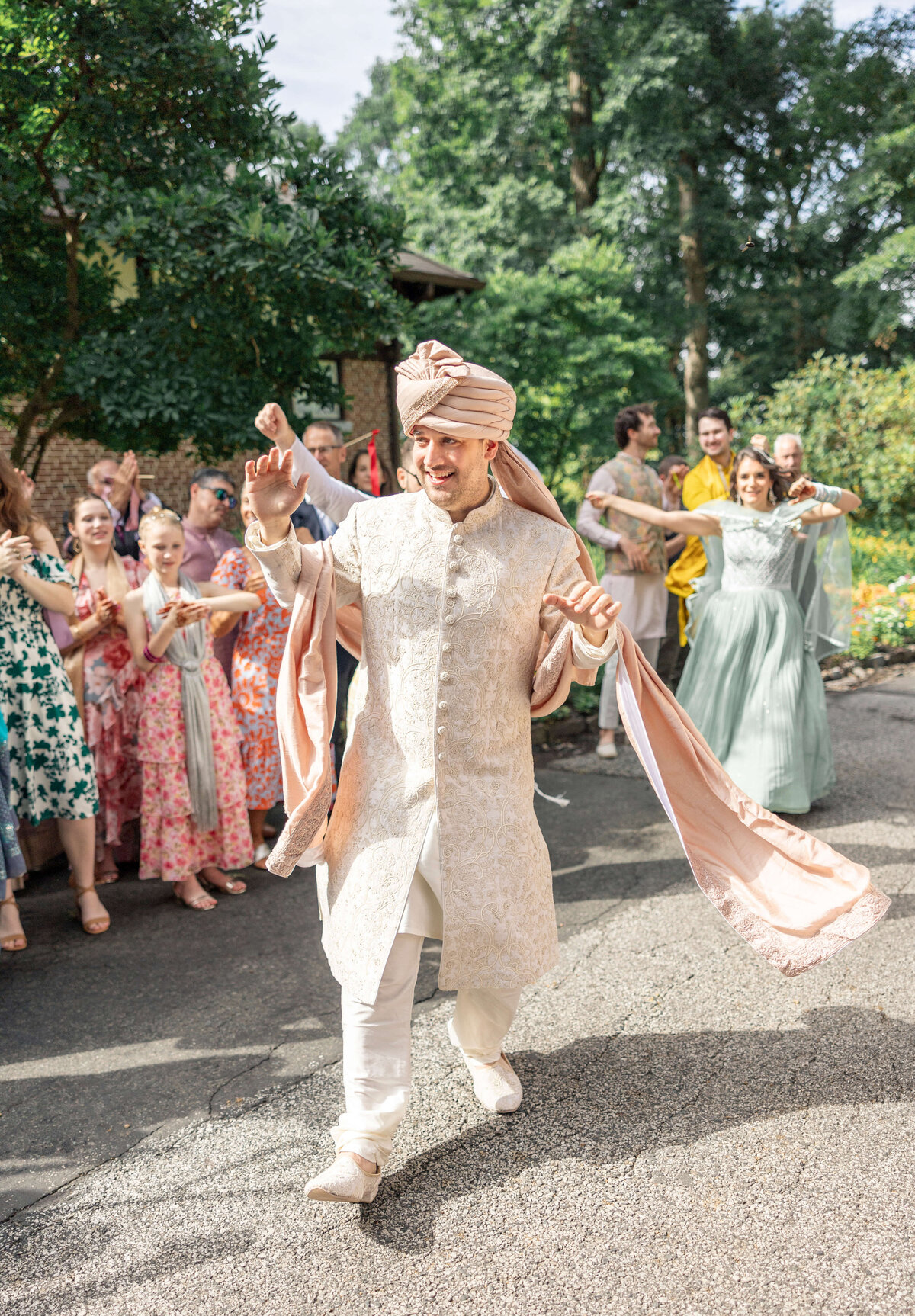 A joyful man in a cream and pink traditional Indian outfit and turban leads a festive outdoor procession. Guests in colorful attire, clapping and dancing, follow him. The scene is lively, set against a backdrop of greenery.