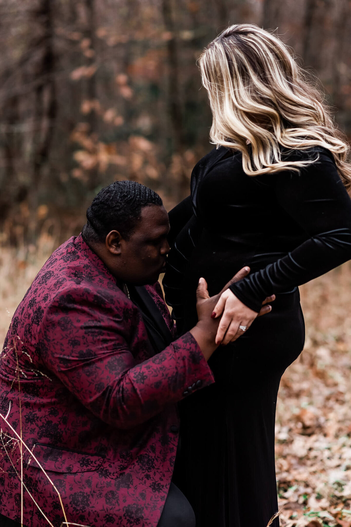 A man wearing a burgundy sports jacket kissing a woman's growing belly