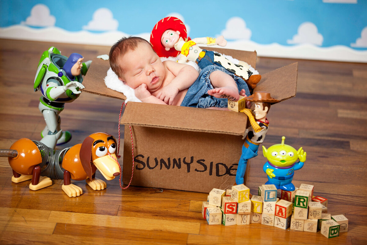 A Jacksonville baby photographer captures a moment as a baby sleeps in a "Sunnyside" box, surrounded by Toy Story characters like Buzz Lightyear and Woody. Nearby, alphabet blocks spell "SKLAR" on the wooden floor, adding charm to this playful scene.