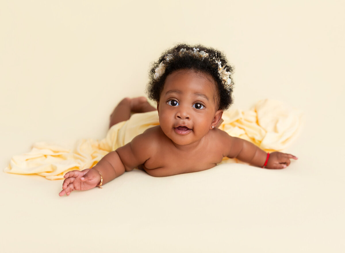 6-month baby girl is lying on her belly for a milestone photoshoot. Baby has her head lifted and is looking at the camera. Baby is wearing a yellow floral crown.