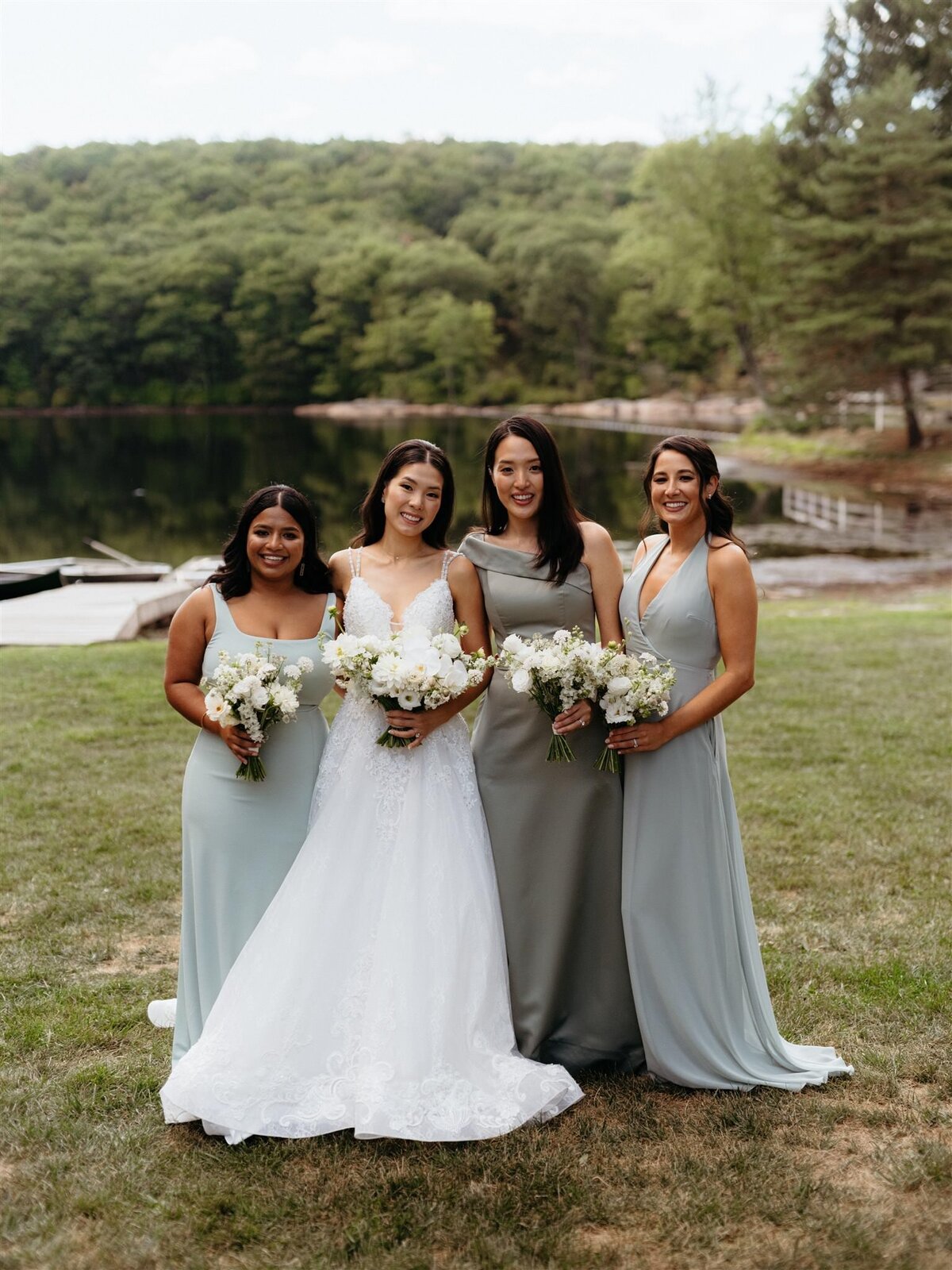 Bride and bridesmaids in floor-length sage green dresses pose on grass in front of lake and trees at Cedar Lakes Estate venue in Catskills