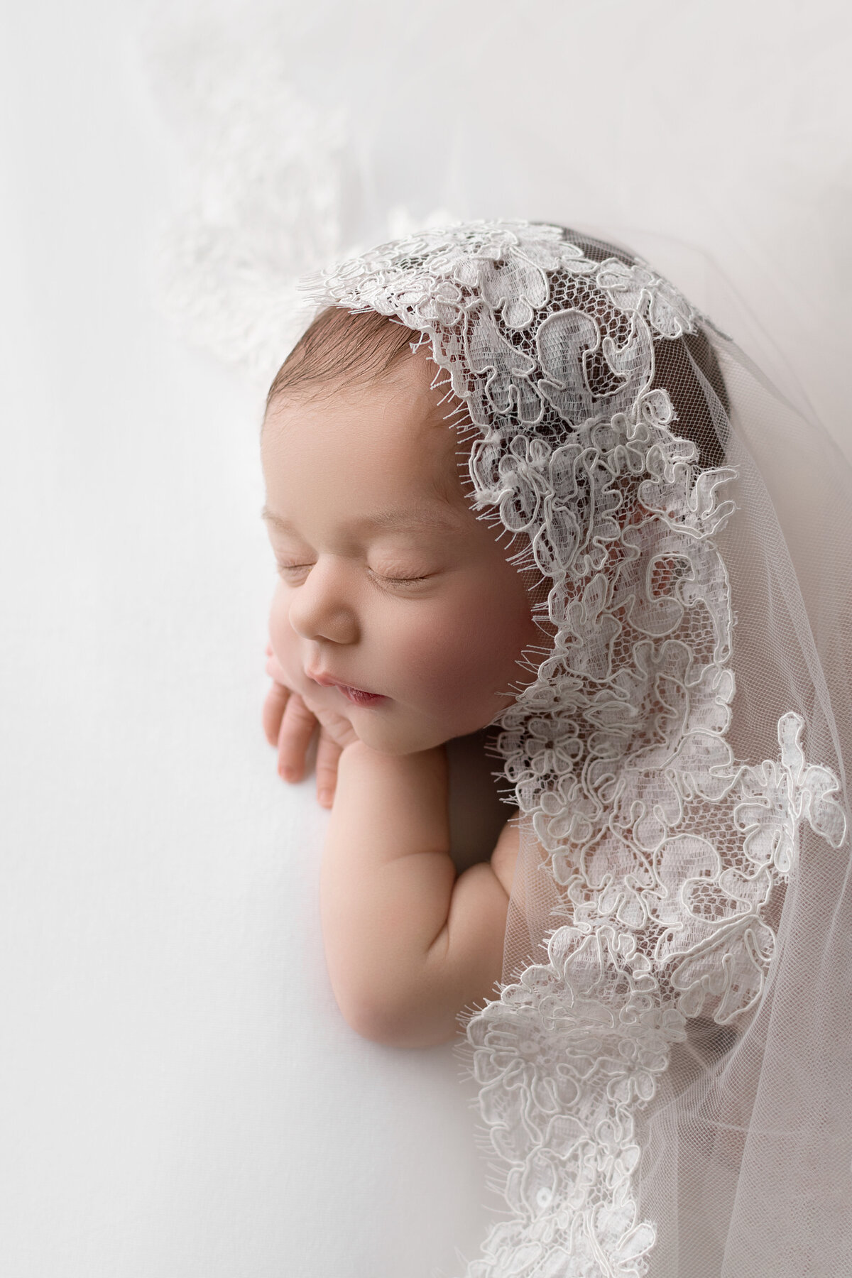 Aerial image. Newborn baby girl is sleeping on her belly with her hands folded under her chin. Her mom's bridal veil is draped overtop of her. Captured by New Jersey's best newborn photographer Katie Marshall.