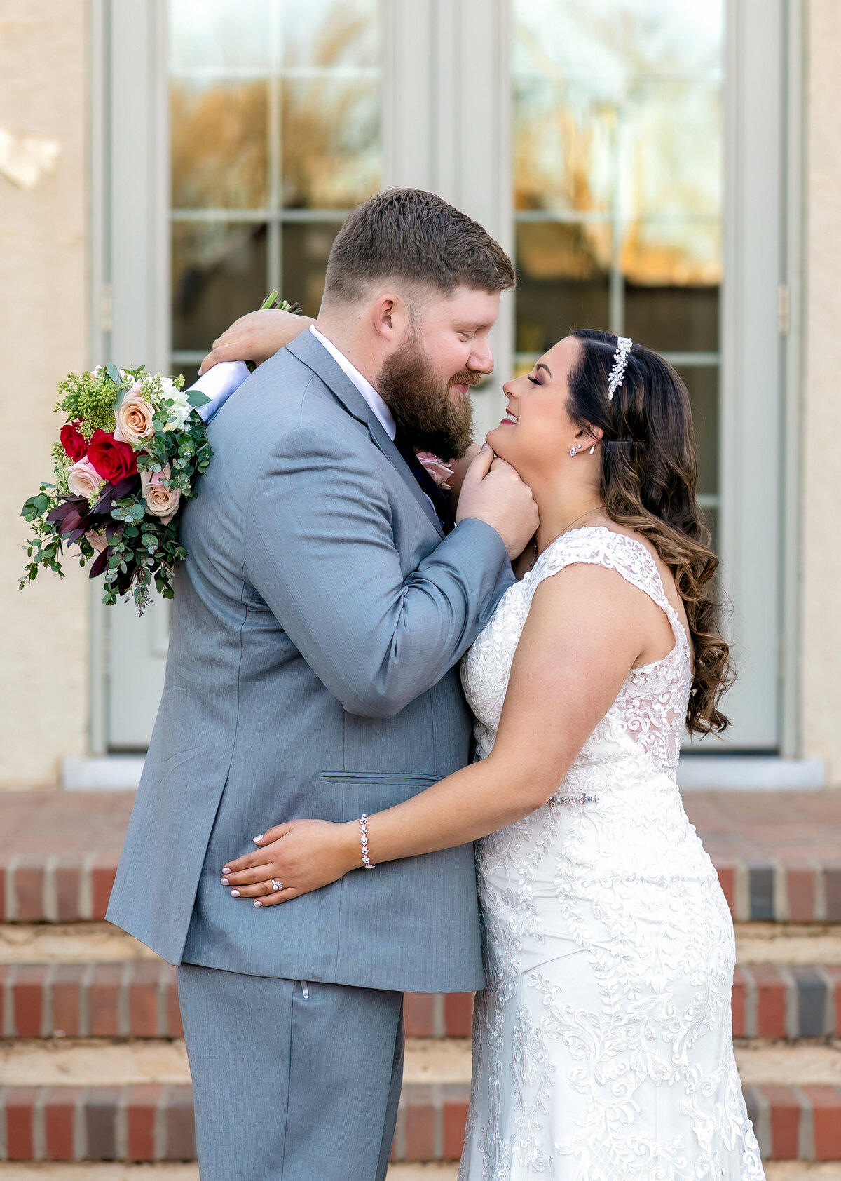 Groom is wearing a gray suit and bride is wearing a lace wedding dress and holding her bouqet over the grooms should while the groom cups her chin to pull the bride in for a kiss at the TTU Merket Alumni Center in Lubbock TX