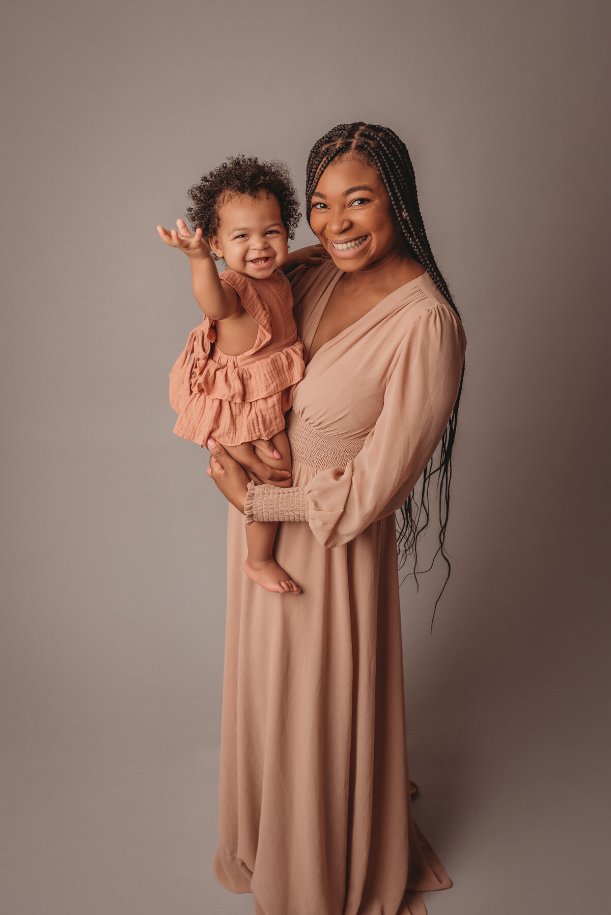 Family portrait on light grey backdrop with mom holding one year old baby girl and they are smiling at camera, wearing nude pink and creams