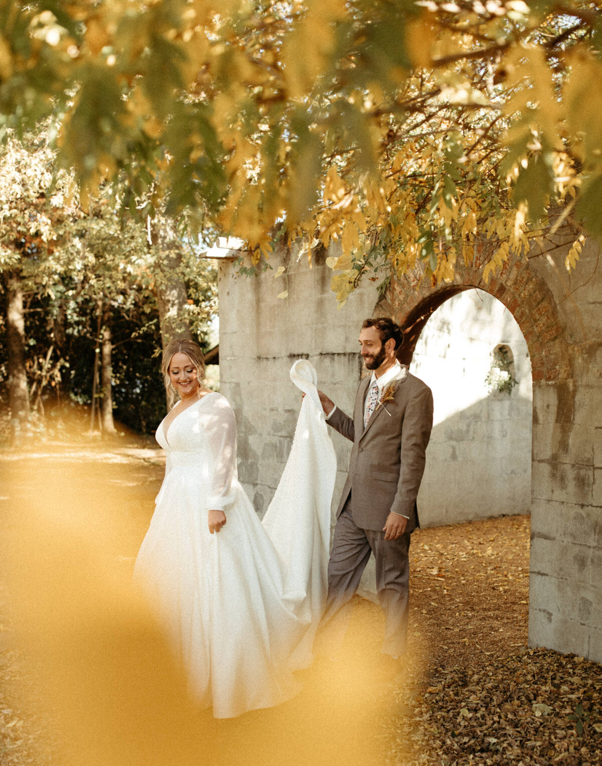 Groom holding bride's wedding dress train up as they walk underneath the branches of a tree with fall foliage