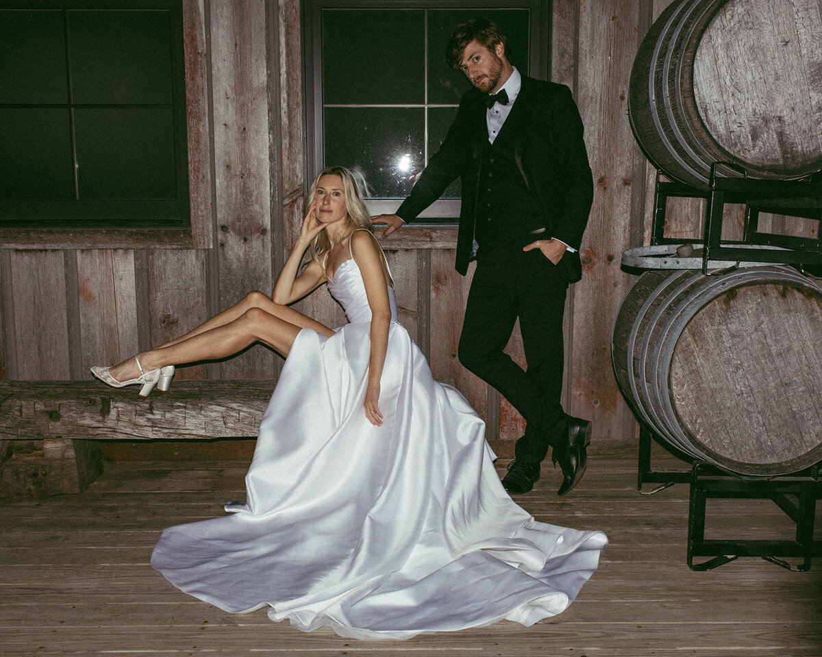 A couple posing elegantly, the woman in a flowing white gown and the man in a black tuxedo, next to wooden barrels in a rustic setting, typical of Iowa weddings.