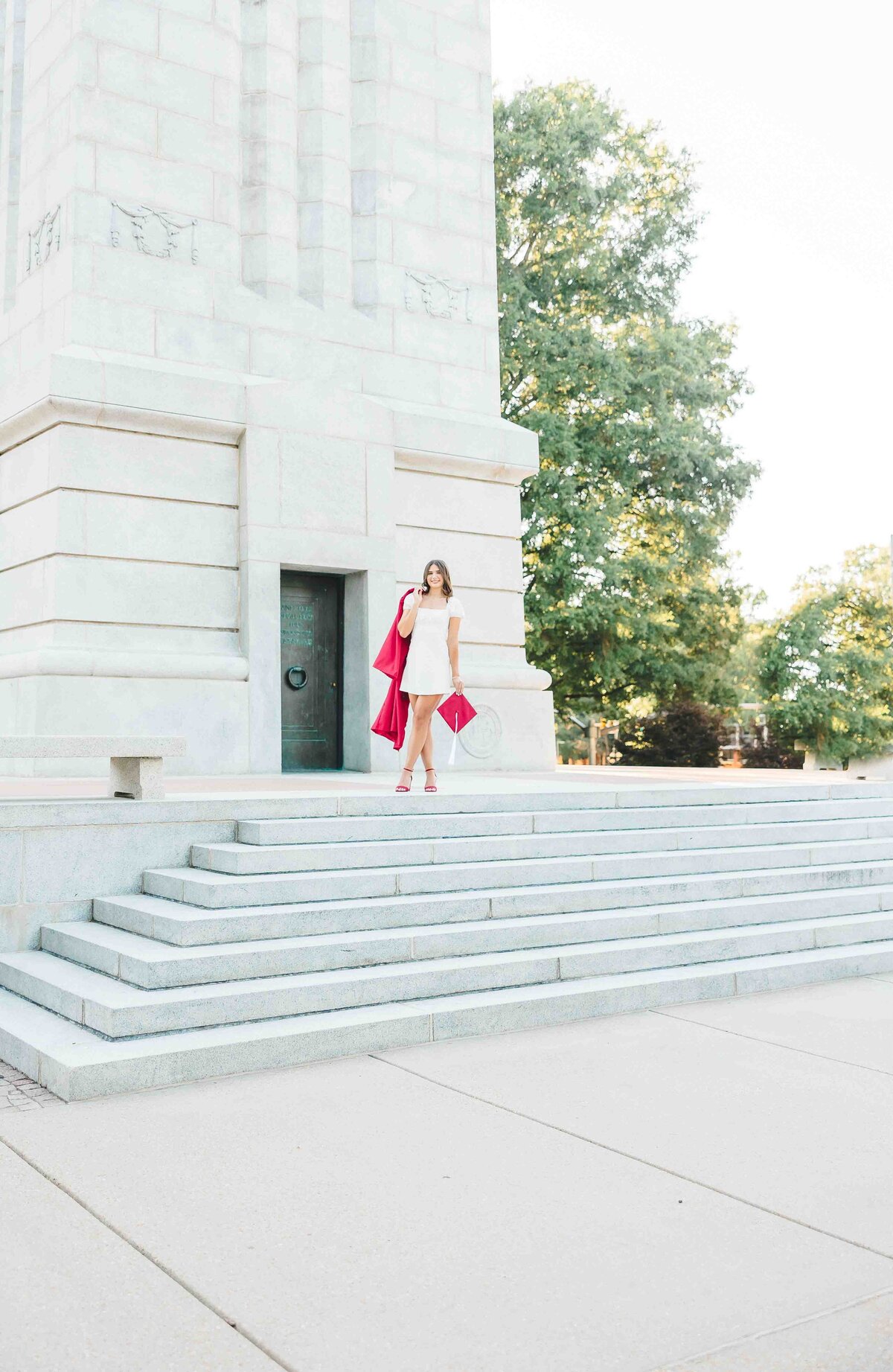 A senior photographer in Fayetteville, NC captures a woman in a white dress on stone steps, holding a red coat and small red purse. She gazes to the side with one hand near her face, framed by a large stone building and lush trees.