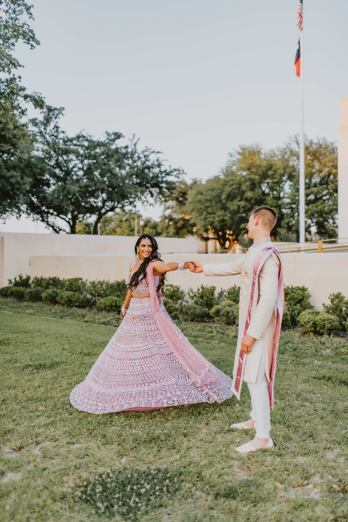 A couple dances gracefully on a lawn. The woman wears a pink lehenga, and the man wears a matching traditional outfit. Both look joyful and elegant.