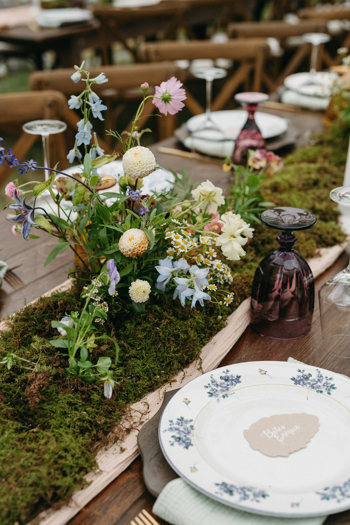 Wildflower-inspired wedding table arrangement featuring dahlias, chamomile, delphinium, and seasonal greenery growing naturally from a moss runner with vintage purple glassware and floral china at an outdoor reception.