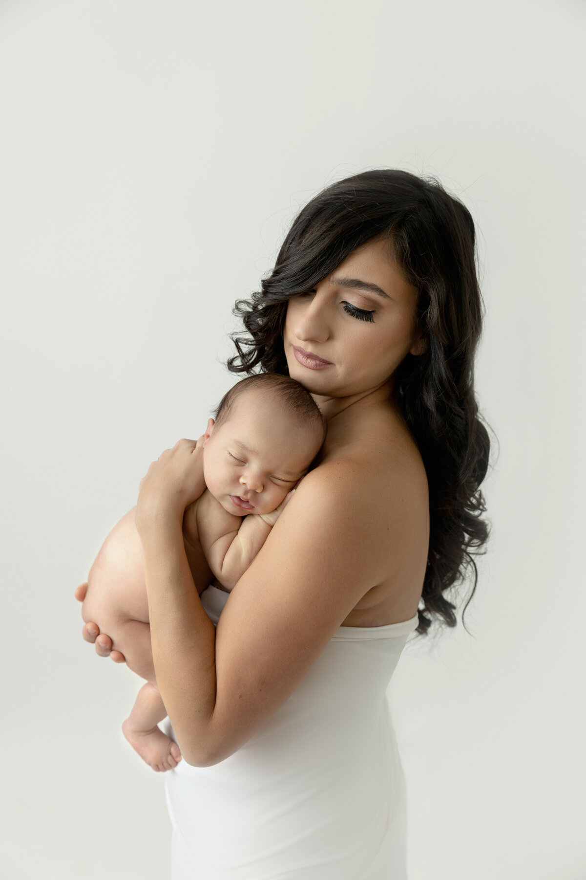 A mother in a white silk dress stands in a studio holding her sleeping newborn baby against her chest