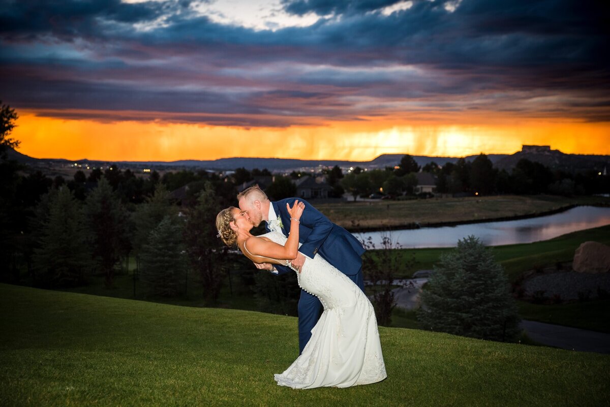 A groom dips his bride for a kiss with a colorful sunset in the background.