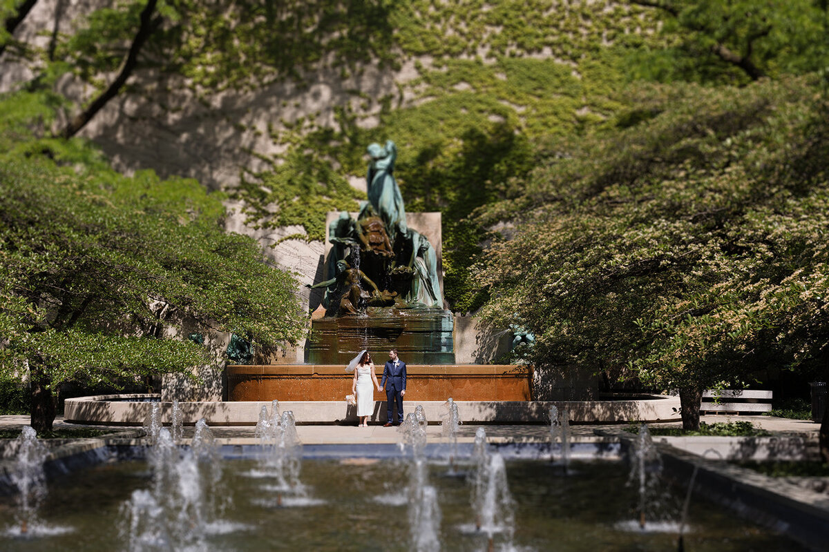 Just Married photo session pulled back photo of couple standing in the Art Institute Garden with the fountain sprays in front of them and the fountain statue and ivy wall behind them.