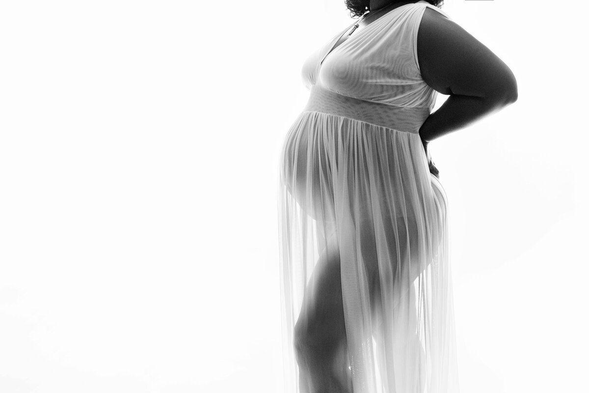 A mother to be stands in a studio in a sheer maternity gown showing her bump