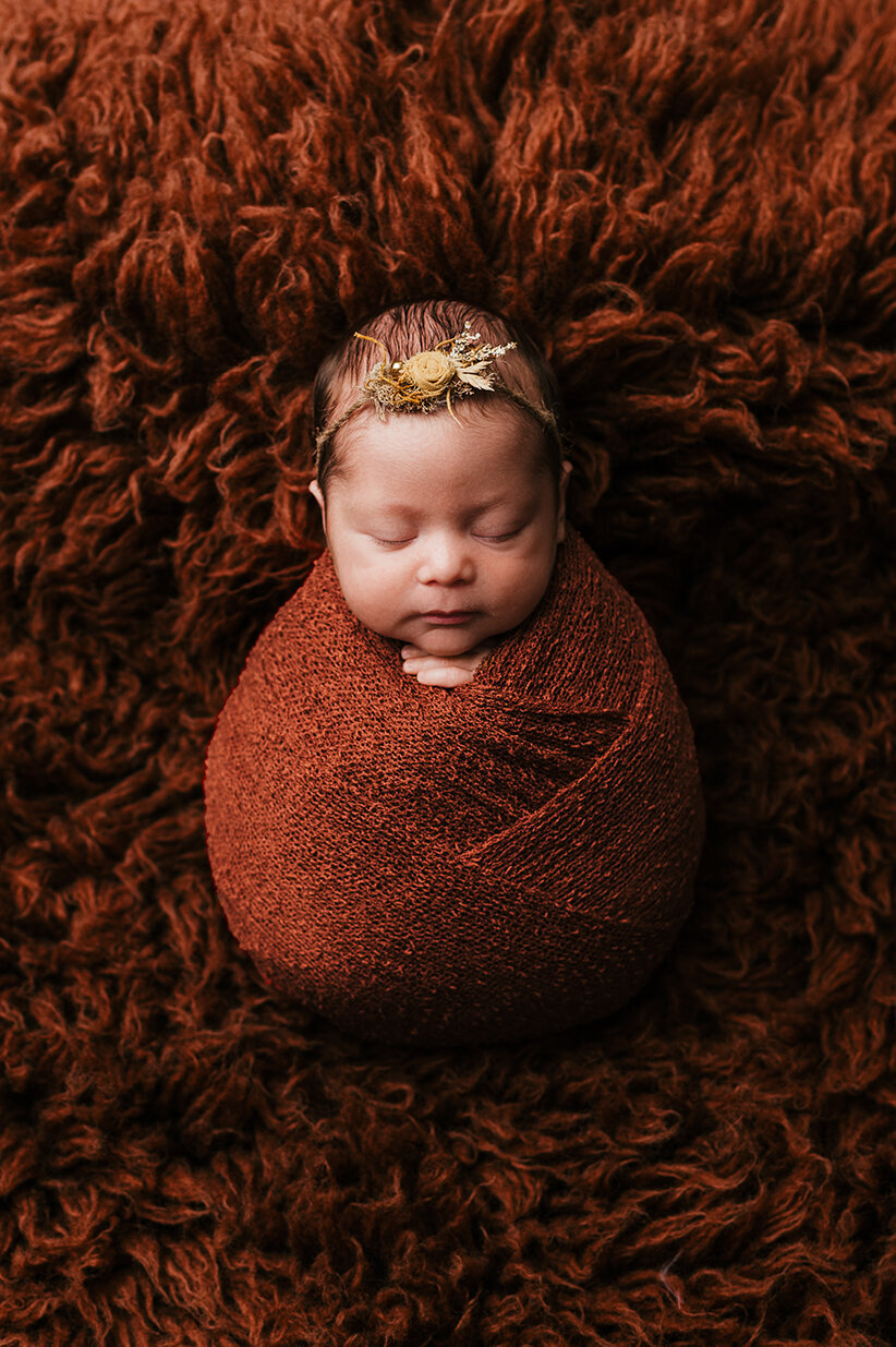 Sleeping baby wrapped in a rust colored wrap laying on a fuzzy rust colored rug