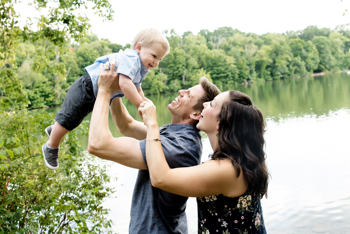 mom and dad swing one year old son in air in front of lake